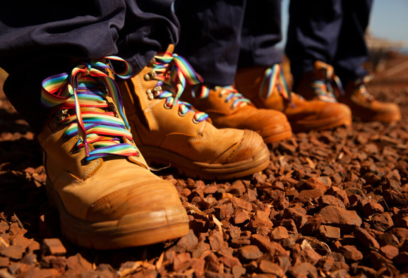 Close-up of a line of work boots with rainbow-colored laces on clay soil