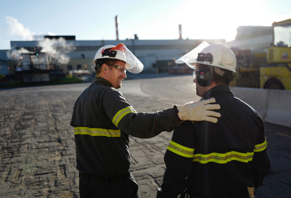 Deux employés de Rio Tinto portant une veste et un casque, devant une usine de traitement – l’un pose sa main sur l’épaule de l’autre